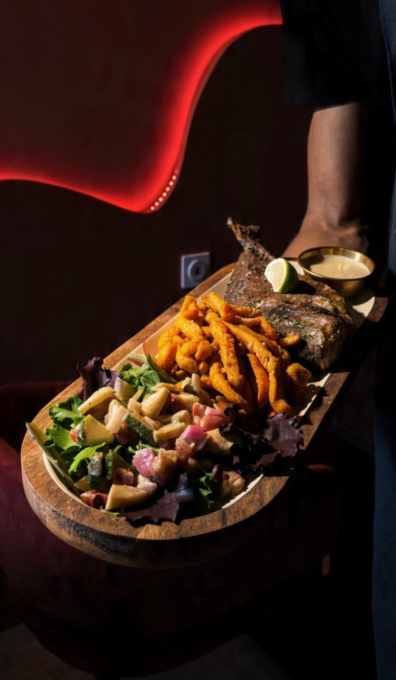Dorade braisée entière, salade de crudités, frites de patate douce et sauce, servie sur une planche en bois — West Afrik Nîmes